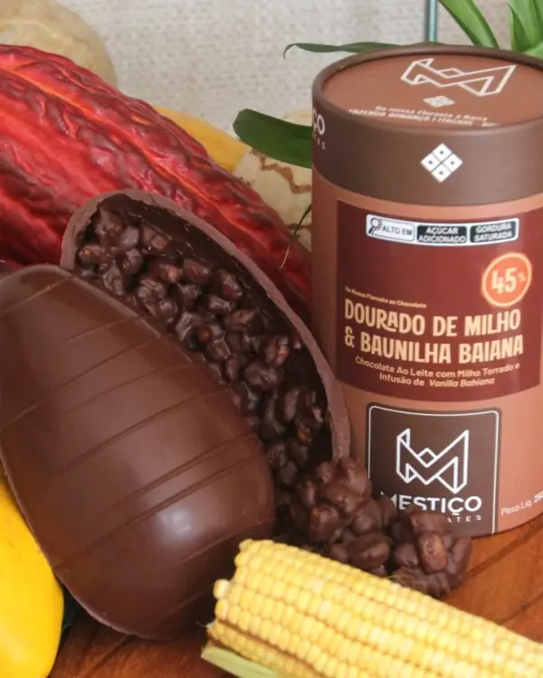 Ovo de Páscoa Mestiço 45% aberto mostrando recheio de milho torrado e baunilha baiana com espiga de milho ao lado.