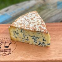 Fatias e cunha do cremoso Queijo Mofo Azul Duzu da BelaFazenda descansando sobre rústica tábua para cortes em sítio.