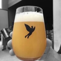 Copo de cerveja com a Melonrise da Trilha, mostrando sua cor amarelo-turva e espuma cremosa, com o logo da cervejaria em destaque.