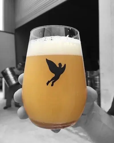 Copo de cerveja com a Melonrise da Trilha, mostrando sua cor amarelo-turva e espuma cremosa, com o logo da cervejaria em destaque. Copo de cerveja com a Melonrise da Trilha, mostrando sua cor amarelo-turva e espuma cremosa, com o logo da cervejaria em destaque.