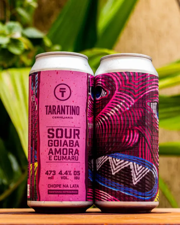 Duas latas da Cerveja Sour Goiaba, Amora e Cumaru da Tarantino, com o rótulo rosa e a arte psicodélica em destaque sobre uma base de madeira.