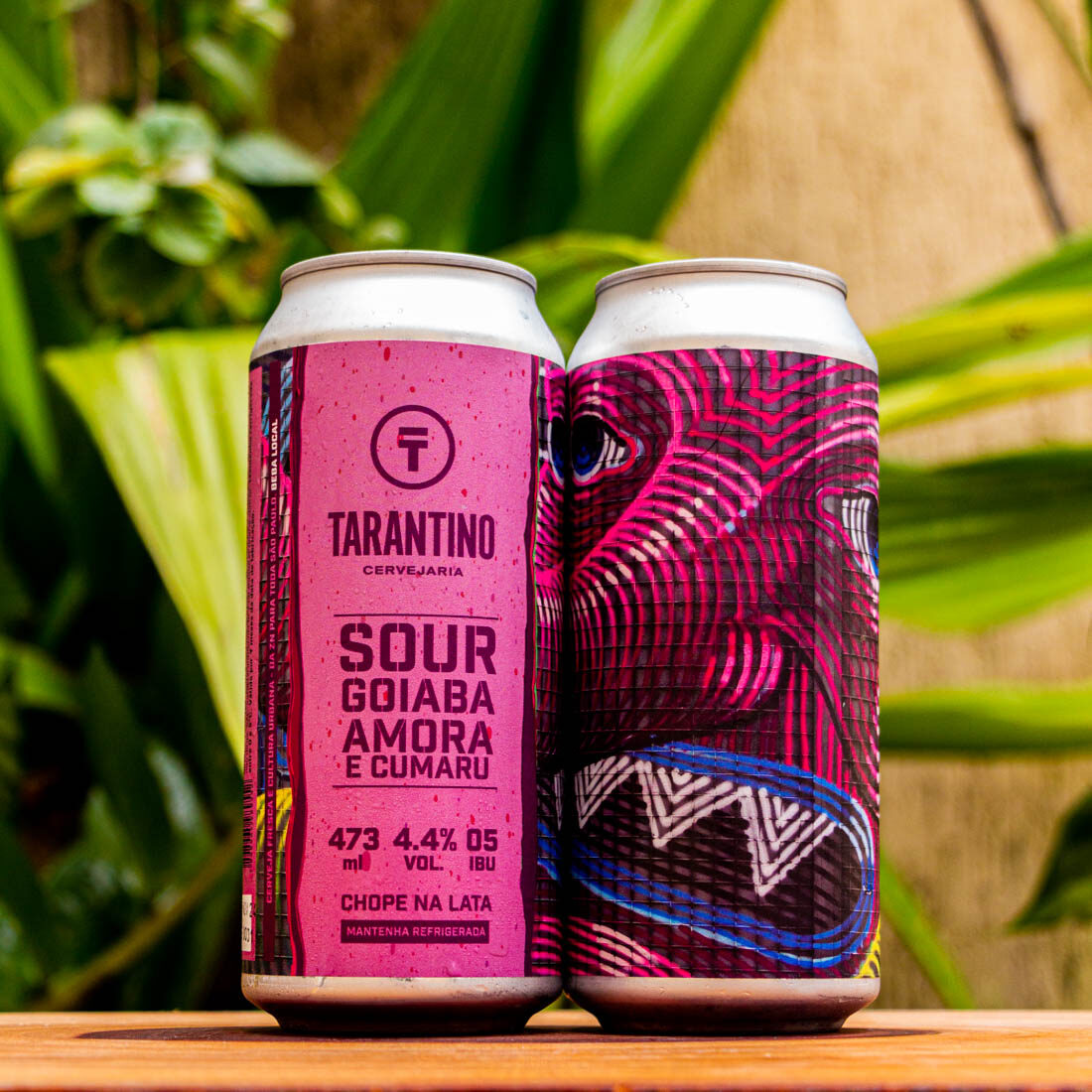 Duas latas da Cerveja Sour Goiaba, Amora e Cumaru da Tarantino, com o rótulo rosa e a arte psicodélica em destaque sobre uma base de madeira.