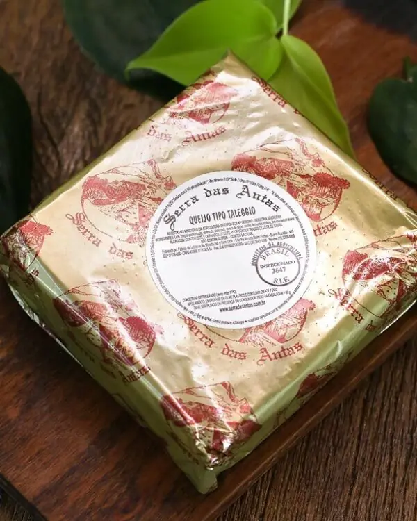 Queijo Taleggio Serra das Antas em embalagem dourada premium sobre tábua de madeira rústica e folhas verdes.