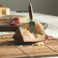 Close-up de uma cunha de Queijo Tulha sendo cortada com faca especial, evidenciando os cristais de tirosina e a massa quebradiça