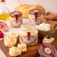Creme de Queijo de Cabra La Popote em pote de vidro aberto, servido com espátula sobre tábua com pães artesanais e tomilhoCreme de Queijo de Cabra La Popote em pote de vidro aberto, servido com espátula sobre tábua com pães artesanais e tomilho