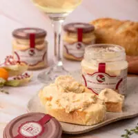 Pote de vidro do Creme de Queijo de Cabra La Popote aberto em cima de tábua rústica com pão artesanal e ervas frescas