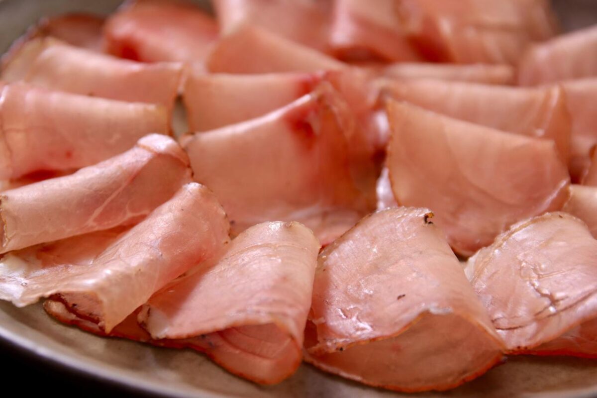 Close-up de fatias finas e rosadas de lonza artesanal dispostas em uma tábua de madeira gourmet com alecrim.