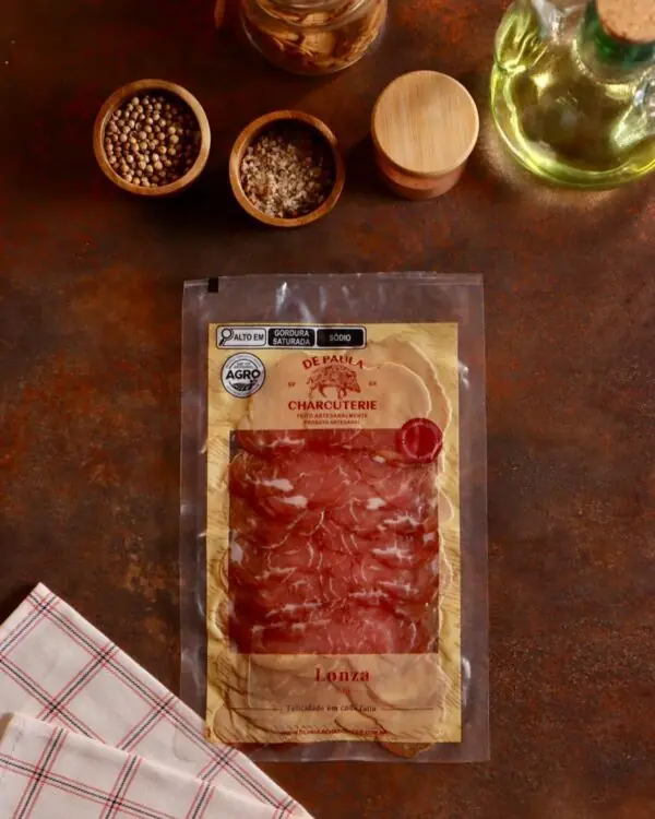 Embalagem de Lonza Maturada De Paula Charcuterie 80g fatiada em cenário gastronômico rústico com azeite.