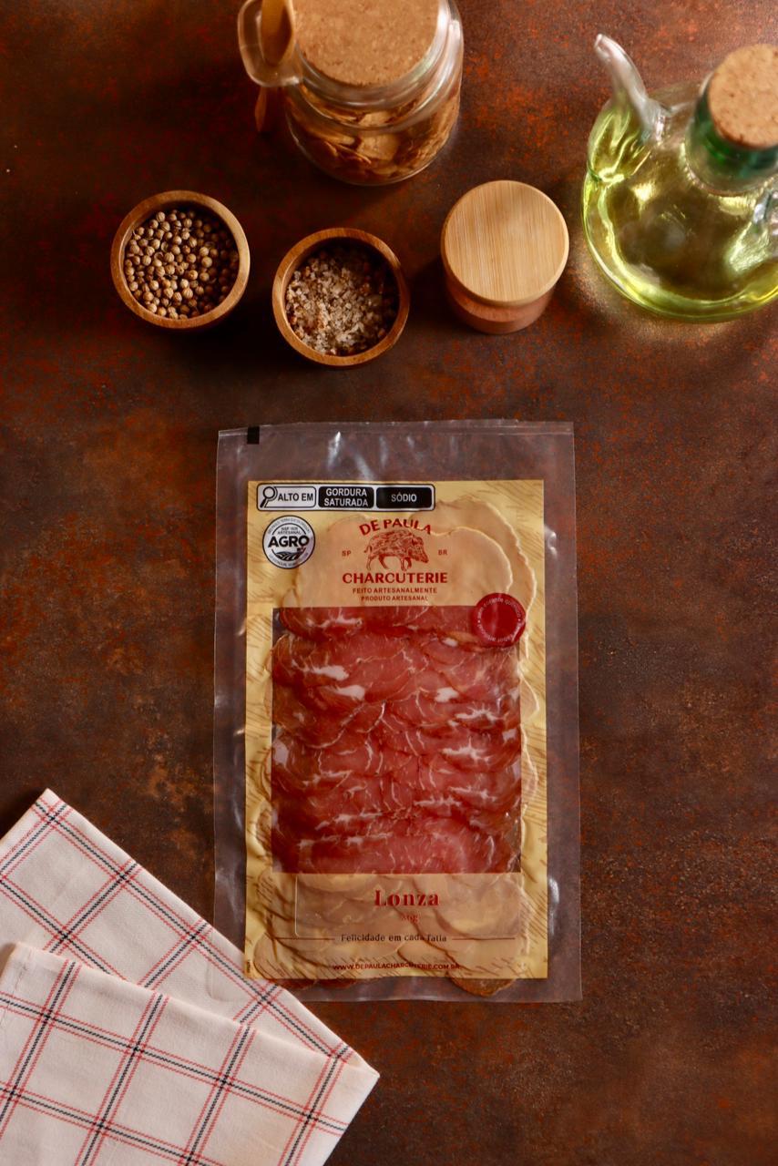 Embalagem de Lonza Maturada De Paula Charcuterie 80g fatiada em cenário gastronômico rústico com azeite.