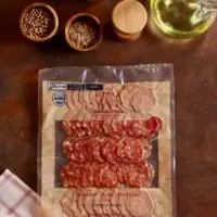 Embalagem de Salame Toscano De Paula Charcuterie 100g fatiado em cenário rústico com pimentas e temperos.