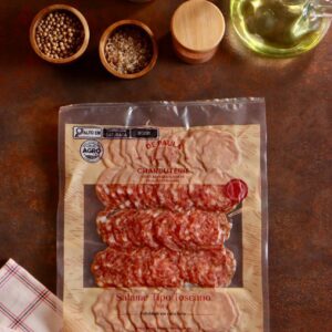 Embalagem de Salame Toscano De Paula Charcuterie 100g fatiado em cenário rústico com pimentas e temperos.