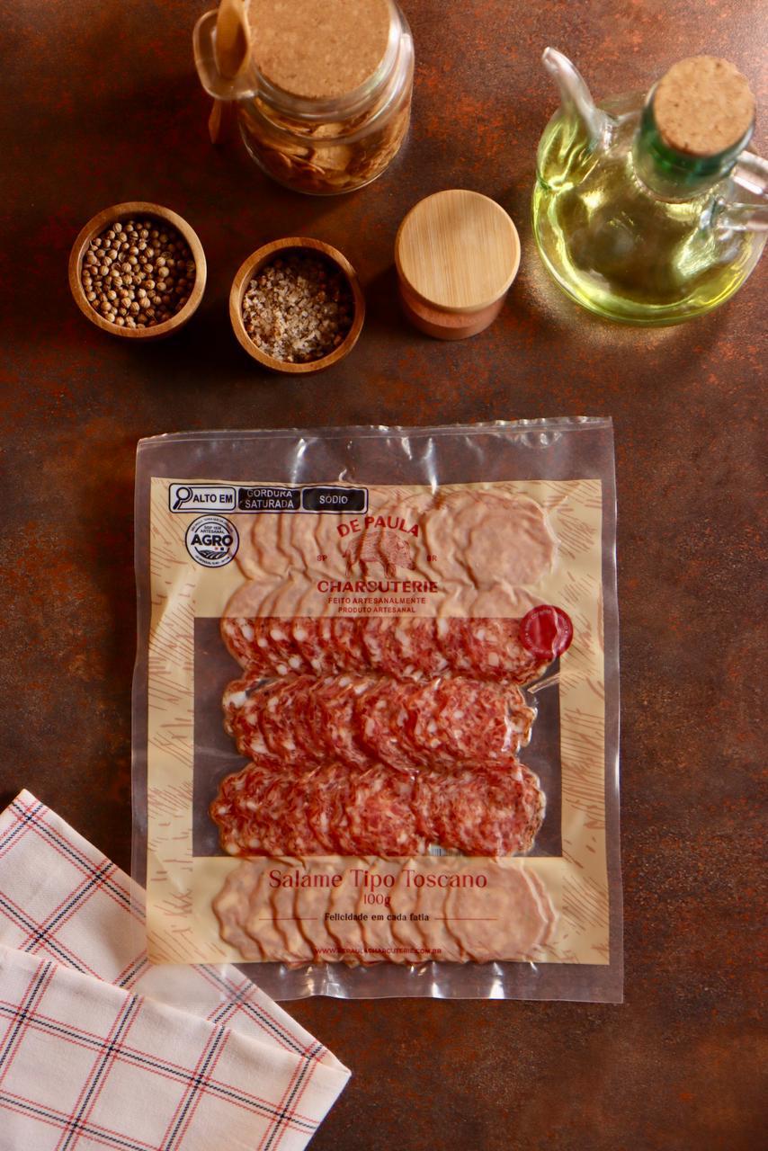 Embalagem de Salame Toscano De Paula Charcuterie 100g fatiado em cenário rústico com pimentas e temperos.
