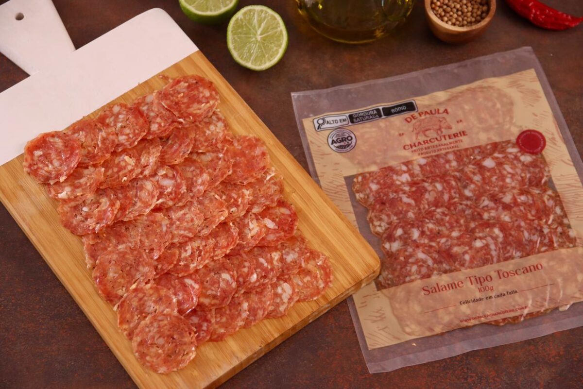 Fatias de Salame Toscano dispostas em tábua de bambu gourmet com limão e alecrim ao fundo.