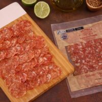 Fatias de Salame Toscano dispostas em tábua de bambu gourmet com limão e alecrim ao fundo.