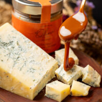 Calda de caramelo de chocolate e laranja sendo despejada sobre queijo gorgonzola em tábua rústica.