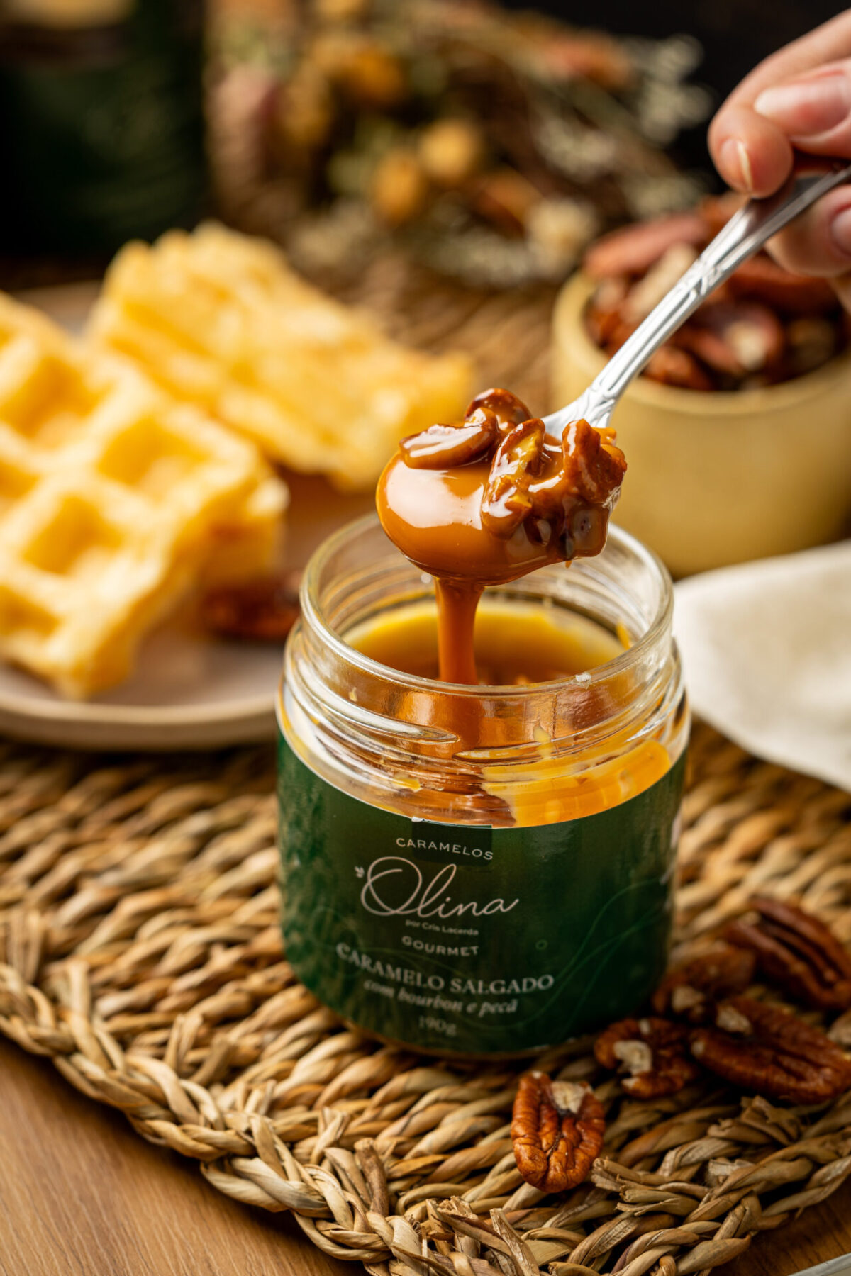 Imagem macro atrativa focando em abastada colher prateada segurando calda densa e grandes castanhas sobre pote de Bourbon Olina ladeando os doces waffles do lanche da tarde agradáveis ao sol e um copo de pécãs puras cruas soltas.