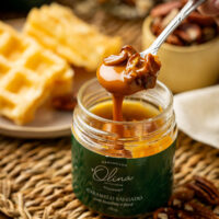 Imagem macro atrativa focando em abastada colher prateada segurando calda densa e grandes castanhas sobre pote de Bourbon Olina ladeando os doces waffles do lanche da tarde agradáveis ao sol e um copo de pécãs puras cruas soltas.