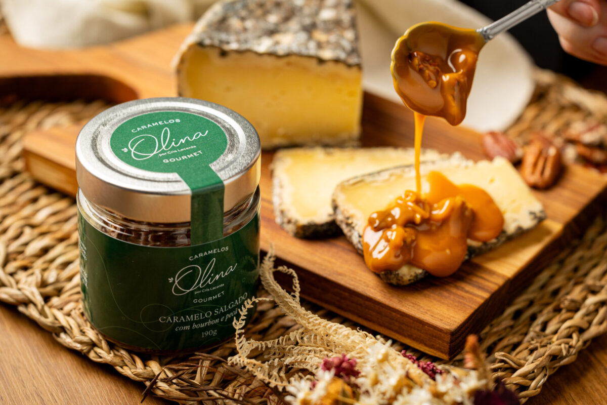 Visão noturna artística sobre Frasco da linha do Rótulo Verde envolto posicionado sobre tábua retangular do queijo meia cura sendo recoberto maciçamente e acompanhado em esteira trançada das flores secadas finas artesanais ao fundo.