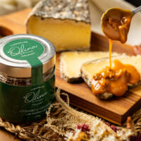 Visão noturna artística sobre Frasco da linha do Rótulo Verde envolto posicionado sobre tábua retangular do queijo meia cura sendo recoberto maciçamente e acompanhado em esteira trançada das flores secadas finas artesanais ao fundo.