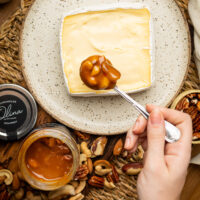 Calda cremosa de caramelo picante e pedaços de nozes caindo devagar sobre fatia rústica de queijo brie macio.