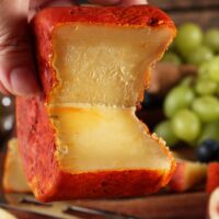 Mão segurando queijo Jordão Pasta Dura partido textura quebradiça casca urucum uvas ao fundo