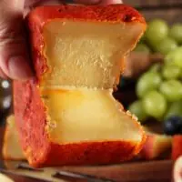 Mão segurando queijo Jordão Pasta Dura partido textura quebradiça casca urucum uvas ao fundo