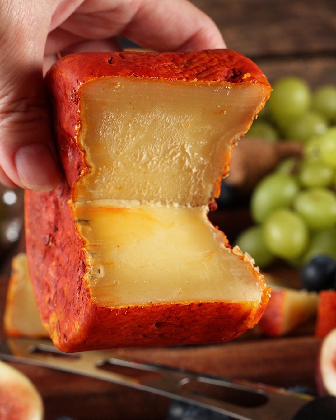 Mão segurando queijo Jordão Pasta Dura partido textura quebradiça casca urucum uvas ao fundo